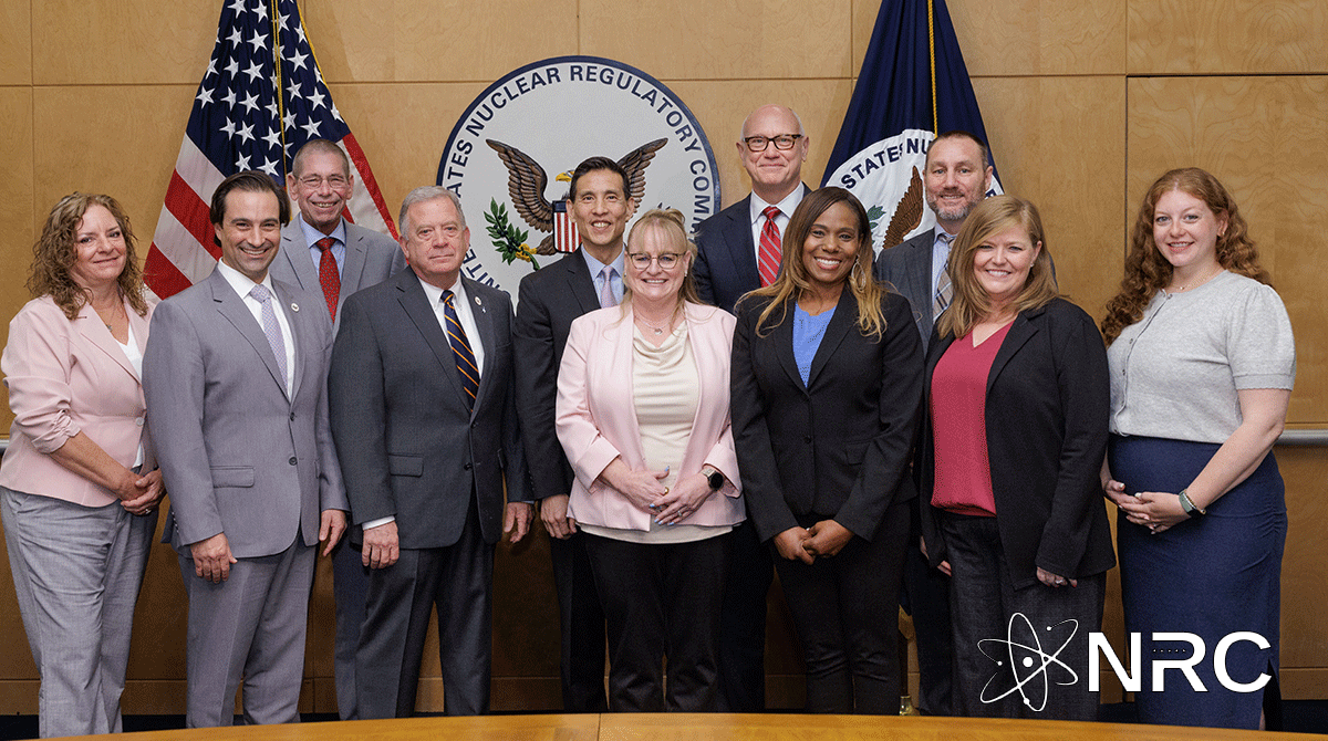 Image is a group photo of 11 people standing in front of the NRC flag, crest, and American flag.
