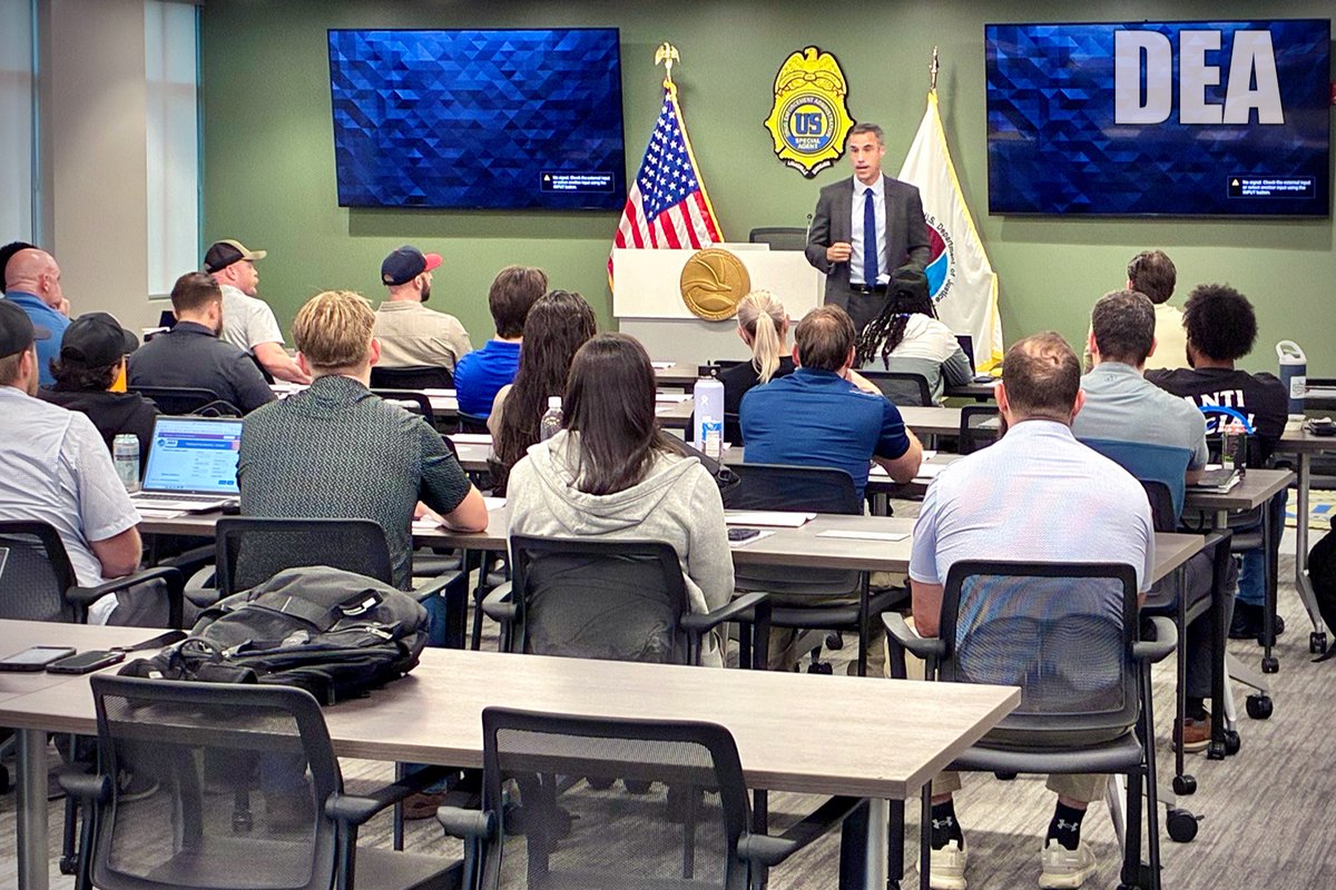 Louisville Field Division Special Agent in Charge Jim Scott speaks to a room full of DEA Special Agents seated at desks with two large displays behind him.