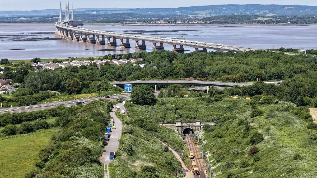 Scenic view of a bridge spanning a wide river, surrounded by lush green landscape. In the foreground, roads and railway tracks lead toward a tunnel.