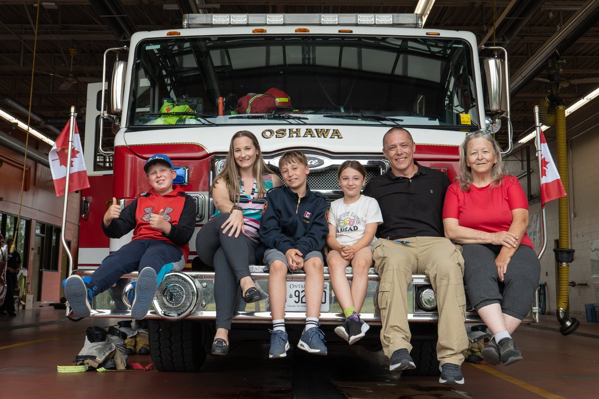 Family sitting on the front bumper of an Oshawa fire truck inside a fire station with Canadian flags on either side.