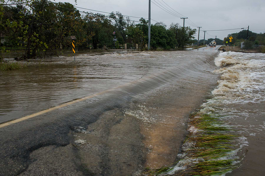 Floodwaters covering a rural road, making it impassable with strong currents visible on both sides.