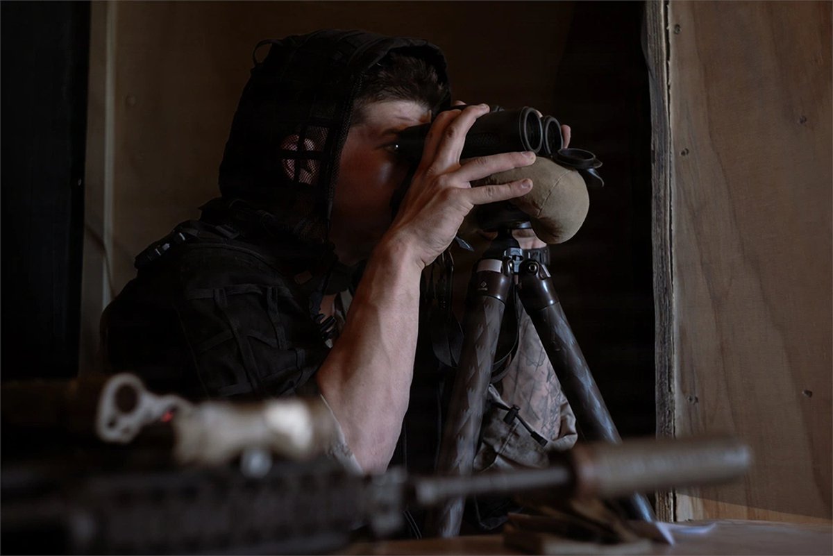 A Marine observes terrain through binoculars from an observation position inside a building.