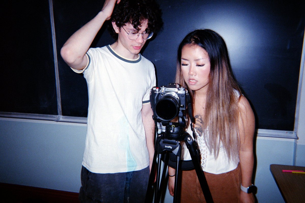 Two people stand in front of a camera on a tripod. One person is touching their head, while the other looks closely at the camera. They are in a room with a chalkboard.