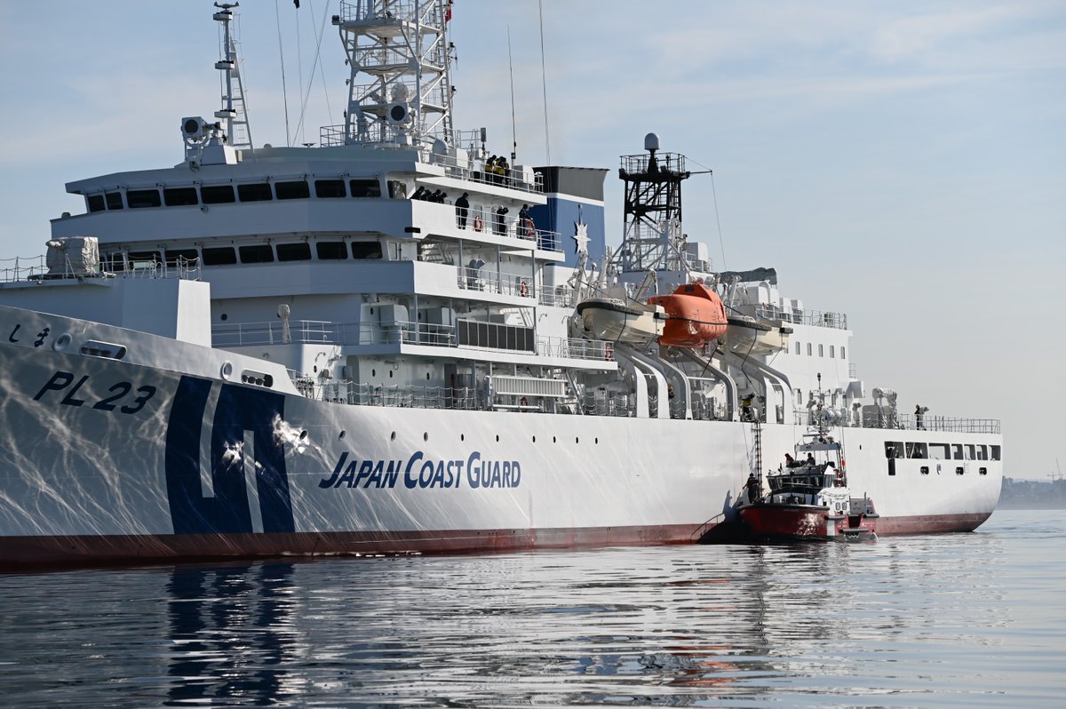 Large Japan Coast Guard white vessel sailing on calm water, accompanied by a small boat nearby, with visible structures and equipment on deck.