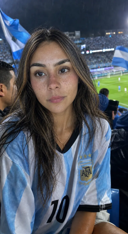 fxabdu01's tweet photo. An intimate straight-on handheld closeup captures a stunning Venezuelan woman seated near the front rows of a packed football stadium during a rainy night matc, wearing an oversized Argentina striped jersey with visible wrinkles, damp fabric texture and a relaxed oversized drape. https://t.co/Qx8OKbY9Gr