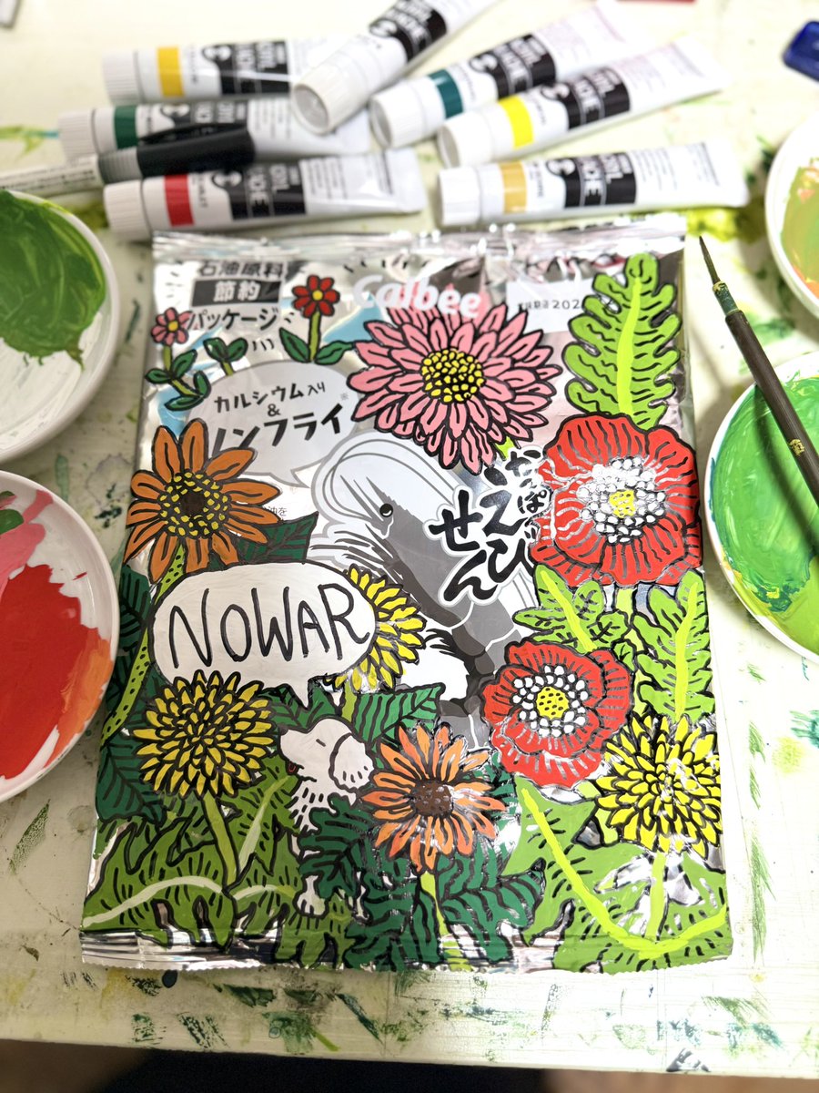 nakamuraippan's tweet photo. "I prefer a colorful world."

Kana Nakamura
2026
Kappa Ebisen bag (petroleum-saving package), acrylic gouache, permanent marker

🌼

I painted this piece on June 25, 2026.
In June 2026, Calbee began releasing products like potato chips and Kappa Ebisen in monochrome printing (petroleum-saving packaging) instead of their usual color printing.

War is a major reason for the shortage of petroleum raw materials.

As of June 2026, due to the escalation of tensions in the Middle East, the blockade of the Strait of Hormuz, and disruptions in logistics, Japan was facing a shortage of imported naphtha (a raw petroleum material).

If there were no war, there would be no oil shortage, and the snacks would have been released in their usual colorful packaging. When I saw the black-and-white bags lined up on the supermarket shelves, I felt the shadow of war gradually creeping into our daily lives.

When this monochrome packaging came out, a certain movement started among illustrators in Japan. It was an attempt to doodle their own drawings using permanent markers (which are also made from petroleum) on the blank spaces left simple by the petroleum-saving design.

Everyone was drawing beautiful pieces, and I thought it was a fun, humor-filled movement. At the same time, however, I felt a certain sense of crisis. I wondered, "Is it really okay to just enjoy and consume this black-and-white packaging caused by an unreasonable war, leaving things as they are? Can we really ignore the underlying problem?"

Petroleum materials are also heavily included in the tools used by painters and illustrators. Not just permanent markers, but all kinds of paints, markers, and even erasers are derived from petroleum. If the oil shortage continues like this, art supplies might become harder to get. If that happens, the next thing to turn monochrome after the packaging might be the very art I create.
I thought to myself, rather than monochrome, "I prefer a colorful world."
So, instead of using a black marker to doodle on the monochrome package and keep it monochrome, I decided to use acrylic gouache (also petroleum-based) to create a colorful drawing.

I usually paint a lot of plants, especially flowers. This is because I love flowers, and since my childhood, plants have always felt closer to me, both mentally and physically, than human beings.
In Japan, when people speak out against war, they are often ridiculed with the phrase "ohanabatake (flower garden) thinking." It is a form of mockery meaning that one is just spouting naive ideals without looking at reality.

"Then, I will paint a flower garden," I thought. If we cannot depict our ideals, it is not true expression.

I surrounded the package with colorful flowers, and to show that we are looking in the same direction as the character in the center, I placed a dog fairy—my alter ego—sitting there, and wrote "NO WAR."

At 10:09 PM on June 25, 2026, I posted the piece "I prefer a colorful world" on X (formerly Twitter). Within a day, the post recorded around 4,000 reposts, 30,000 likes, and over 1.8 million impressions.

What made me personally very happy was that the painter Yoshitomo Nara liked and reposted it from his official X account. This is because I have always been deeply encouraged by Mr. Nara's work, which has long infused the spirit of NO WAR and NO NUKES.

Even now, this painting is being interpreted in many different ways by many people.
The consumption cycle of social media is fast, and this painting will probably be forgotten soon. However, I want to continue my creative practice without ever forgetting that I made a stand here.