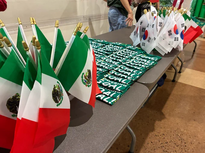 Flags of Mexico and South Korea are displayed on a table. Folded green t-shirts with white text spelling "Qatar FIFA World Cup 2022" are placed between the flags.