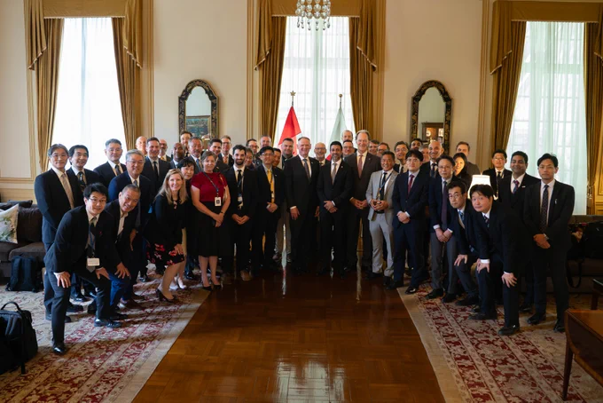 Minister Sidhu and a large group of people in business attire gathered indoors for a formal group photo in an elegant room with tall windows, chandeliers, and decorative mirrors; Canadian and Japanese flags are visible at the center behind the group.