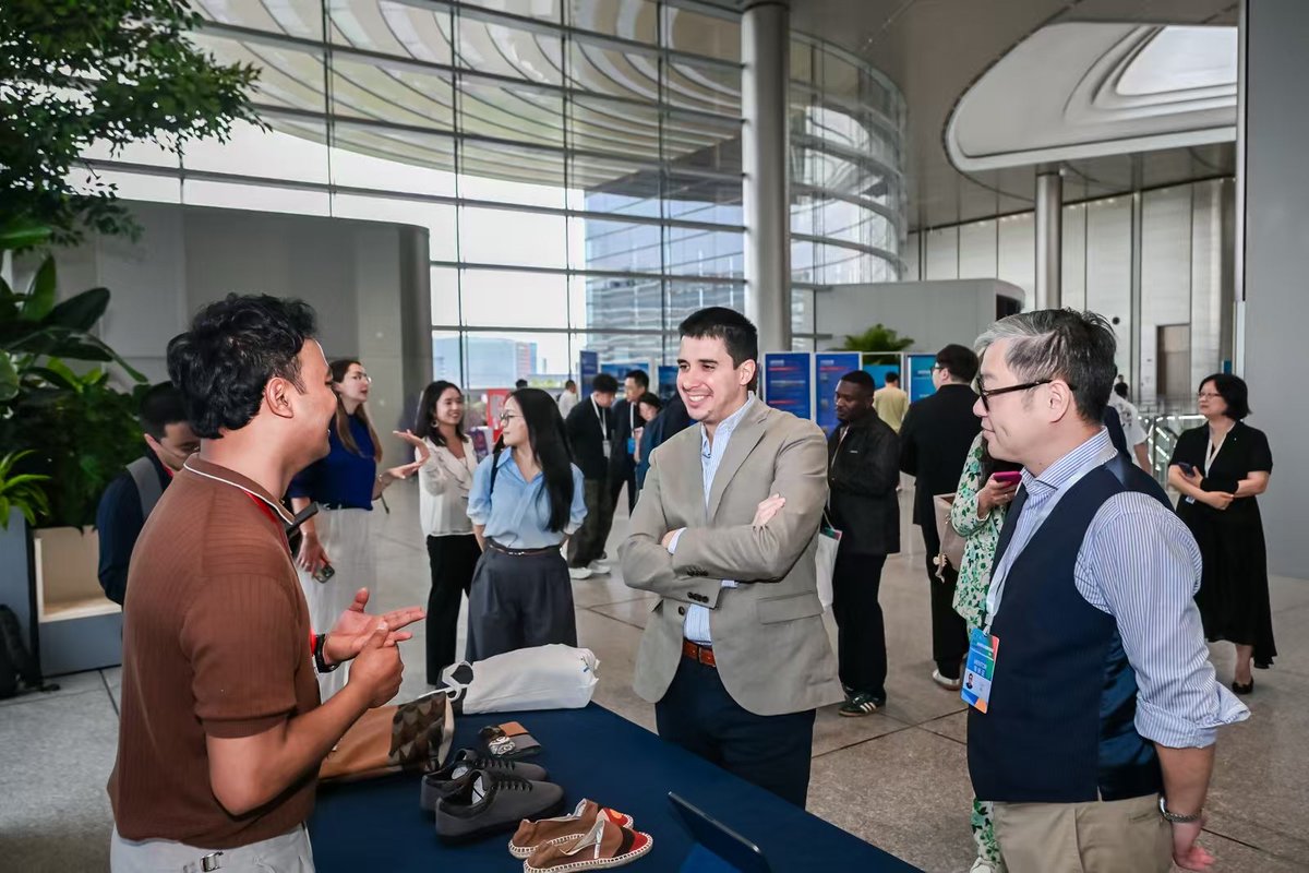 Participants network during the Acceleration Week for Global Youth Development in Shanghai. Several attendees stand in conversation near a display table, with others speaking in the background.