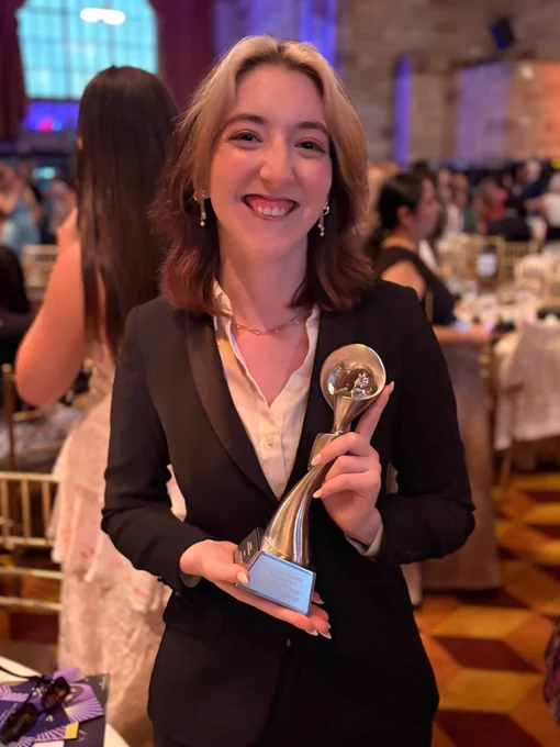 A smiling person in a black suit holds a shiny silver award at a formal event. Blurred, seated guests and elegant decor are visible in the background.