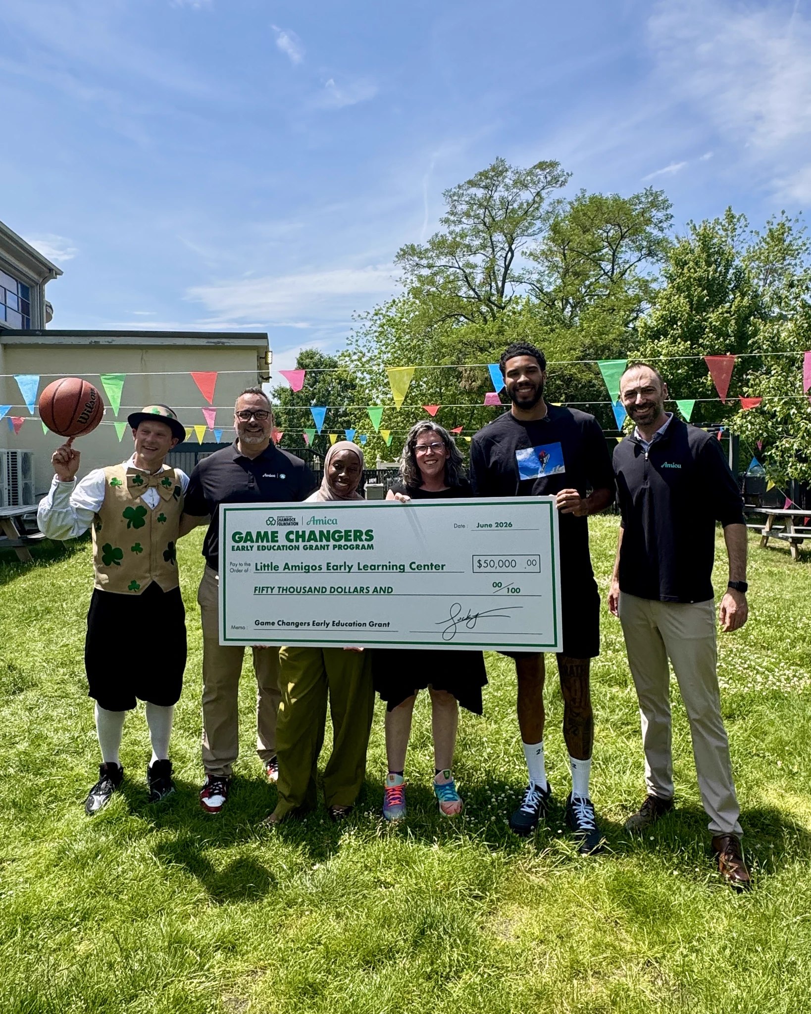 Six people, including a man in a shamrock vest spinning a basketball, pose outdoors with a large check. The check reads 'GAME CHANGERS EARLY EDUCATION GRANT PROGRAM' for '$50,000' to 'Little Amigos Early Learning Center' from the 'Boston Celtics Shamrock Foundation and Amica'. Colorful flags and trees are in the background.