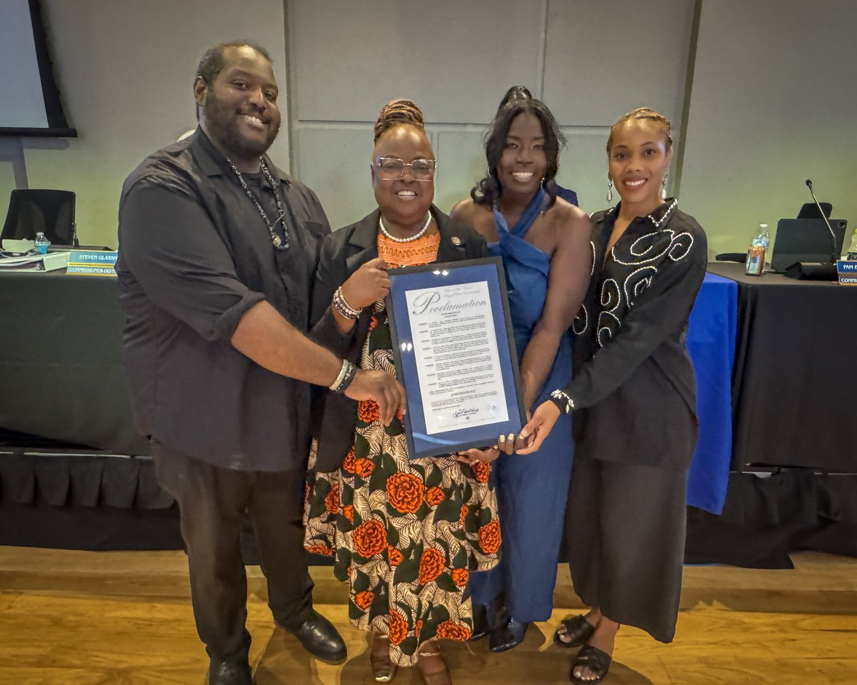 Four people standing together indoors, holding a framed proclamation, dressed in semi-formal attire.