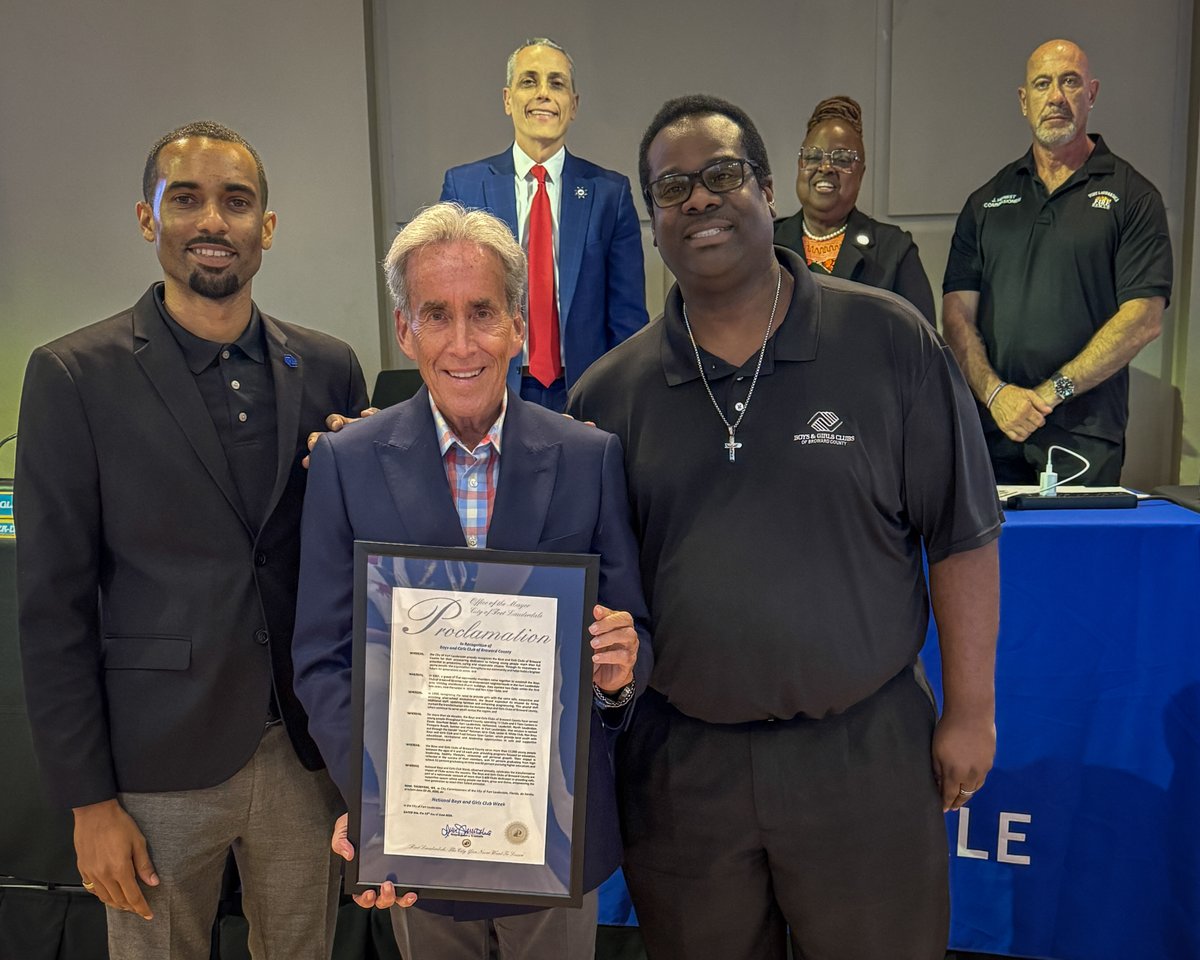 Group of six people posing indoors, one holding a framed proclamation document in front of a blue table with a microphone.