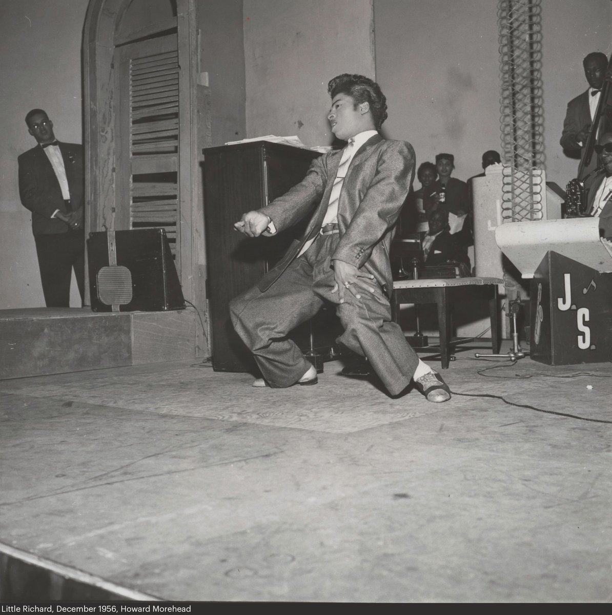 Black-and-white photo of a young suited up Little Richard performing on stage. The band's logo behind him says J.S.