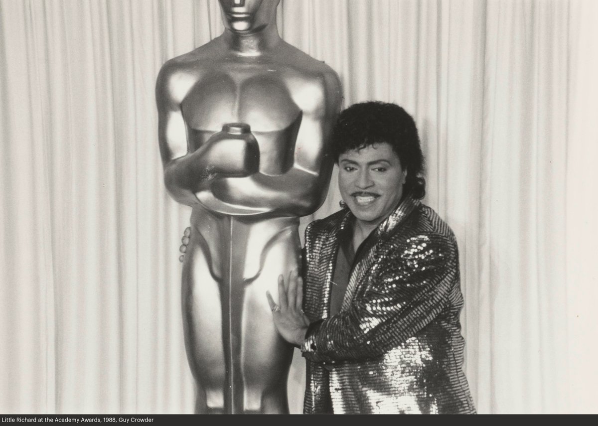 Black-and-white photo of Little Richard embracing an oversized Oscars statuette. He wears a sequined jacket.