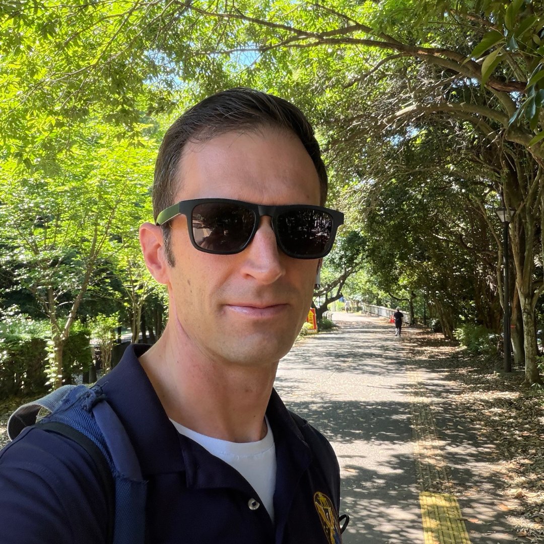 A selfie of a man wearing sunglasses outdoors on a path lined with trees.