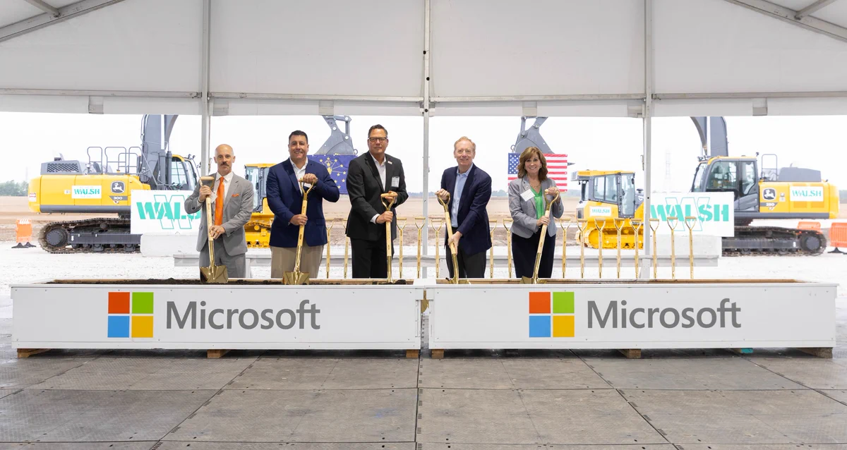 People stand in a row inside a large construction tent, each holding a ceremonial shovel at a groundbreaking event. Behind them are construction vehicles and signs, and in front are barriers displaying the Microsoft logo.