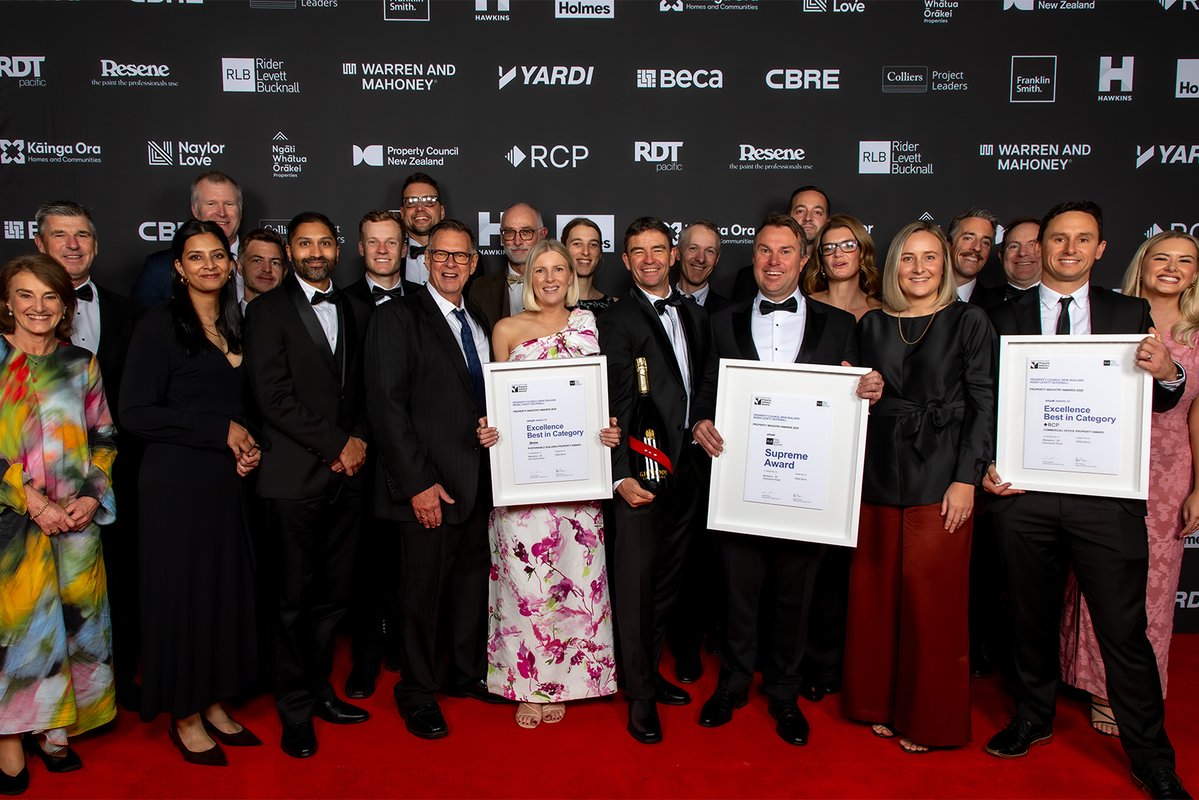 A group of people in formal attire are posing on a red carpet. Three individuals in front are holding framed certificates and a bottle. The backdrop displays logos, including Yardi, Beca, CBRE, Resene, and Warren and Mahoney.