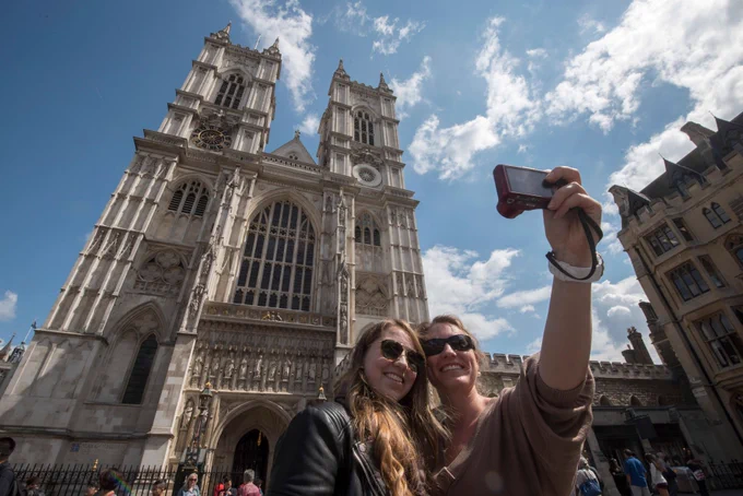 Two women smiling and taking a selfie in front of the grand facade of Westminster Abbey under a blue sky with scattered clouds. The Abbey features intricate carvings, large arched windows, and twin towers.