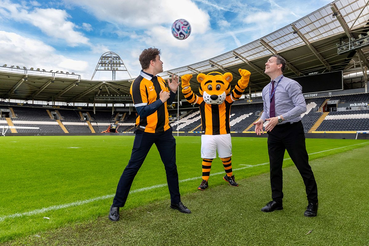 Two people and the Hull City mascot with a ball in a stadium.