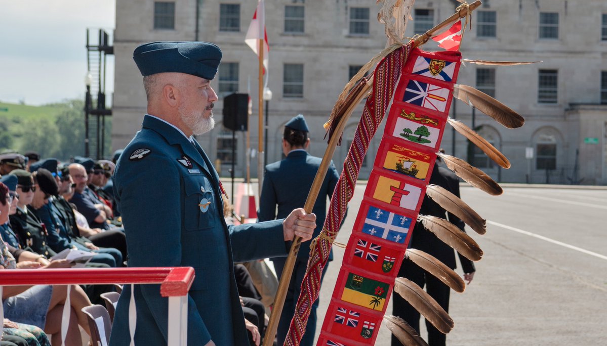 A uniformed military member holds a traditional staff adorned with multiple flags and eagle feathers during an outdoor ceremony. 