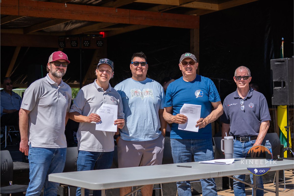 Five participants pose for a photo at Kineepik Métis Local’s Pinehouse Elders Gathering. Two individuals hold signed documents while standing with three others behind a table. A circular logo in the lower-right corner reads “KINEEPIK Métis Local” and features a stylized landscape with trees and an infinity symbol.