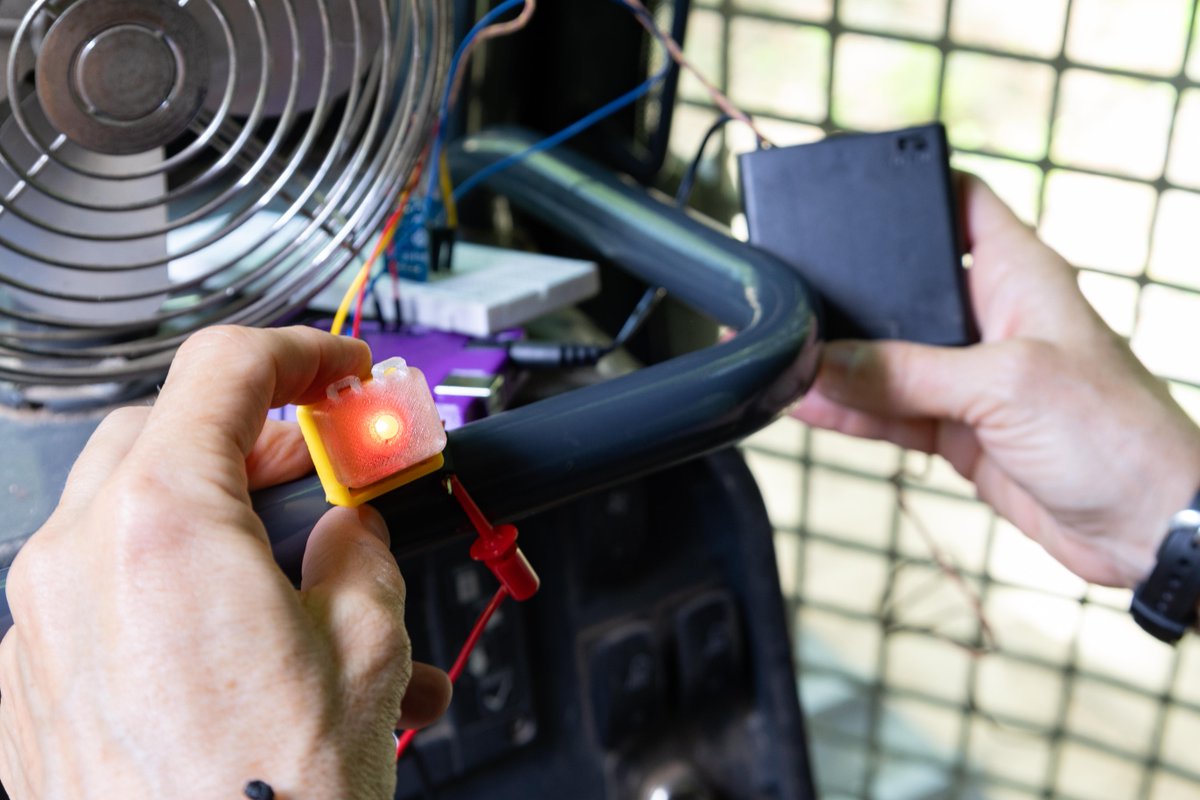 Close-up of hands holding a lit LED indicator inside a fire dozer. The light is connected to a thermal sensor mounted on the fire dozer window. Credit: NASA