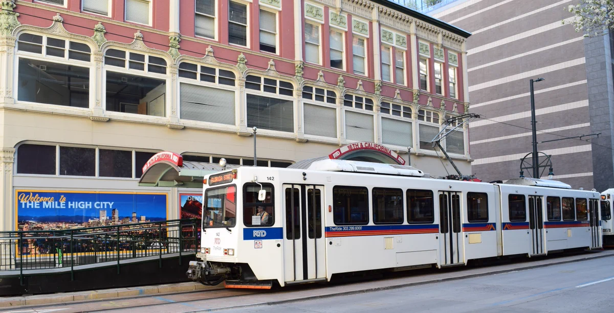 An RTD light rail train pulls into the station at 16th and California streets in downtown Denver. On the building next to the train is a poster that reads "Welcome to the Mile High City."