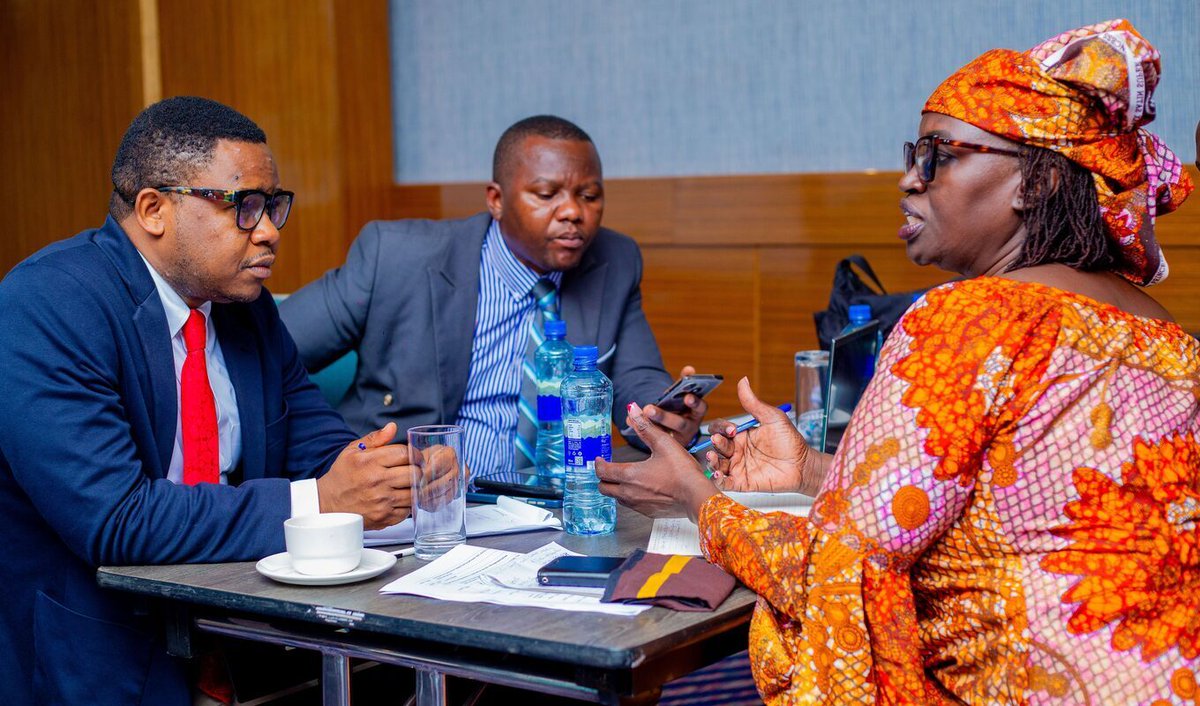 Three individuals in business attire sit at a small conference table, engaged in discussion. Papers and a water bottle are on the table. The setting appears to be a professional meeting.