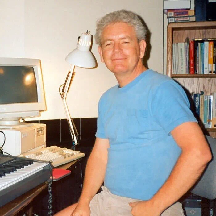 A middle-aged man with gray hair sits beside a desktop computer, wearing a blue T-shirt and beige shorts. He has his left arm resting on a keyboard. The room features a desk lamp and shelves filled with books.