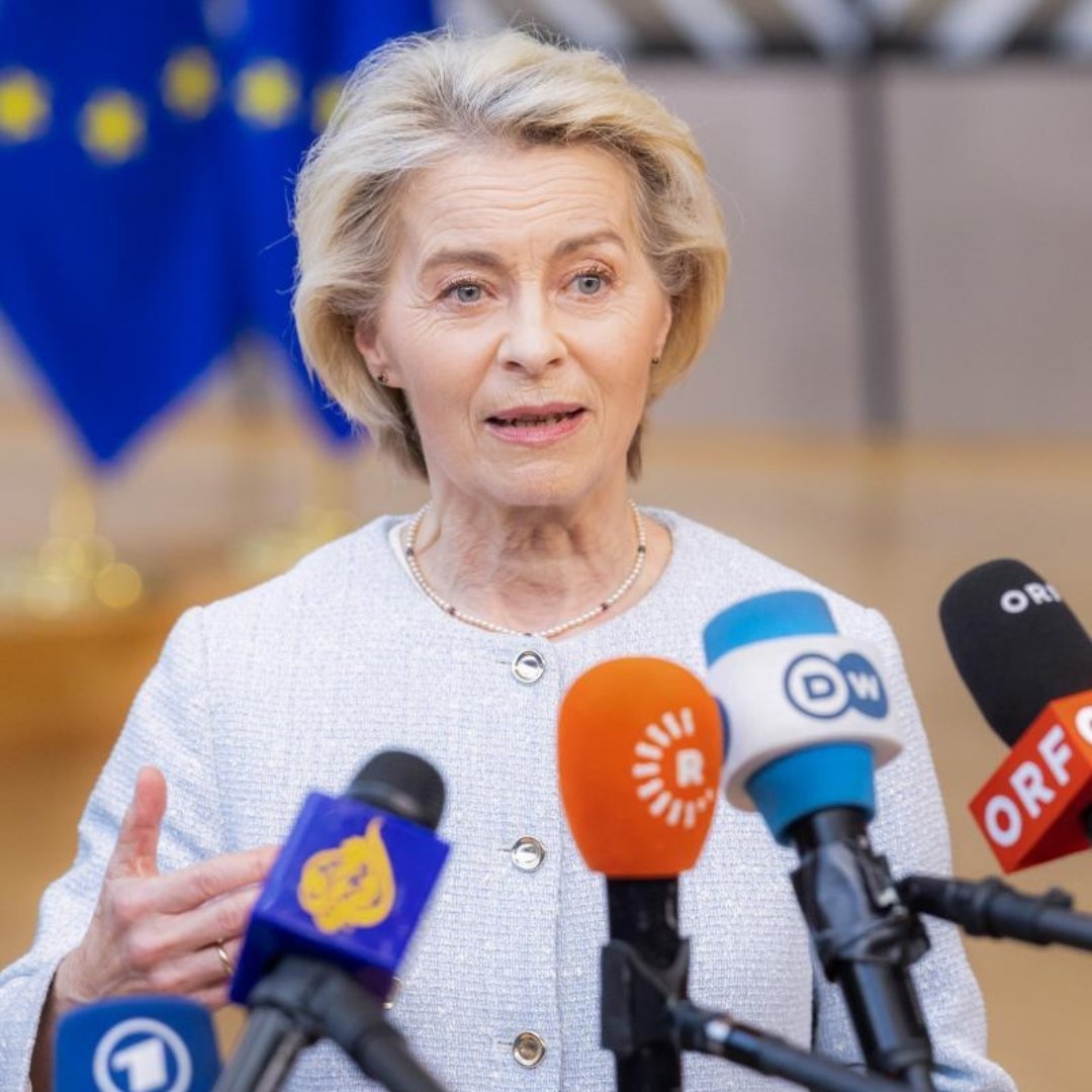 A close-up shot of Ursula von der Leyen speaking directly to a cluster of press microphones, gesturing slightly with her right hand. The microphones feature logos from news organizations like Al Jazeera, DW, and ORF, with a blurred European Union flag in the background. 