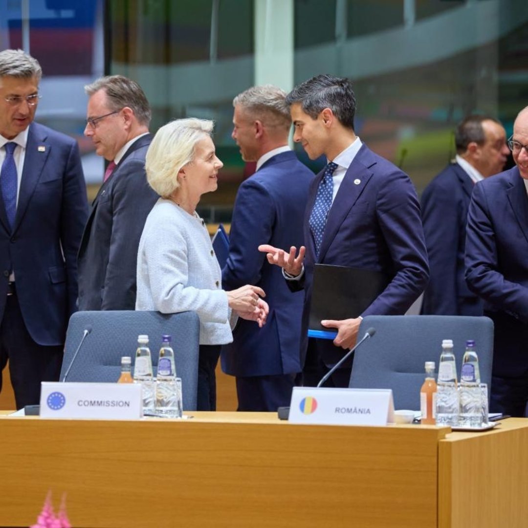 Ursula von der Leyen stands behind a desk and smiles while conversing with Dutch Prime Minister Rob Jetten, who holds a black folder.  