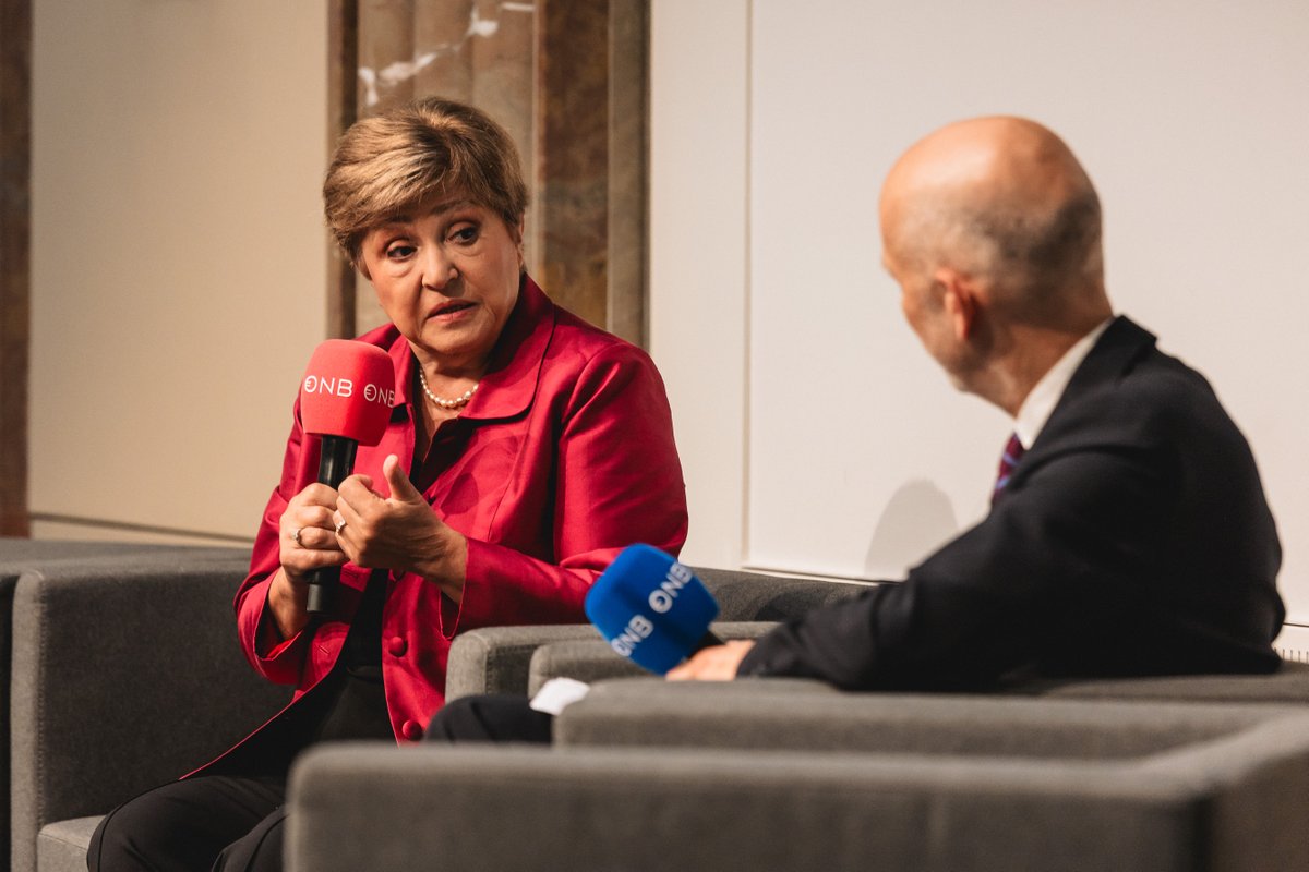 A woman in a red coat speaking into a microphone, facing an older man with gray hair in a suit.