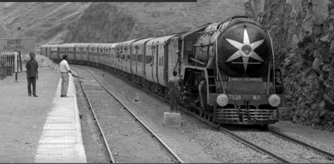 Ananth_IRAS's tweet photo. A beautiful old photo from 1974 of the Koyna Express from Miraj to Bombay VT at Adarki station, hauled by WP 7528, captured by Paul Claxton! The WP engine was built by Canadian Locomotive Co., Kingston, Ontario, Canada - the 102nd engine in a batch of 120 locomotives of this class manufactured under the Colombo Plan Aid Programme in 1955-56.

The Pune–Miraj rail line was originally built by the Southern Maratha Railway (SMR) as an MG line between 1887 and 1890, opening it for traffic in November 1890. As passenger demand and freight increased, the line was converted to BG. Because the original straight alignment featured gradients too steep for heavier BG trains, the iconic Daundaj Horseshoe Curve (a sweeping 180-degree curve) was introduced to ease the ascent and descent through the rocky terrain. #IndianRailways #History