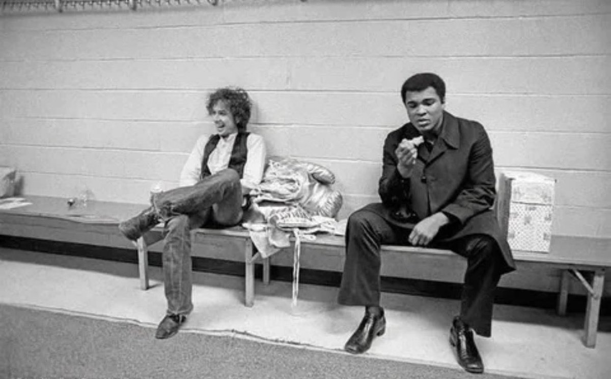 HistoryLBT's tweet photo. Muhammad Ali and Bob Dylan backstage at Madison Square Garden, December 8, 1975 — photographed by Ken Regan, the sole official photographer of Dylan’s Rolling Thunder Revue tour. The giant boxing glove between them on the bench was Ali’s gift to Dylan that evening. The Night of the Hurricane was the closing concert of the tour, a benefit for Rubin Carter — a middleweight boxer Dylan believed had been wrongfully imprisoned for murder, the subject of his song ‘Hurricane.’ Joan Baez, Joni Mitchell, Allen Ginsberg, and Sam Shepard were all part of the tour. Ali tried to turn the evening into a political rally; Dylan responded by dedicating a song to someone who, he noted pointedly, was ‘not running for President.’ Rubin Carter’s convictions were finally set aside ten years later, in 1985.