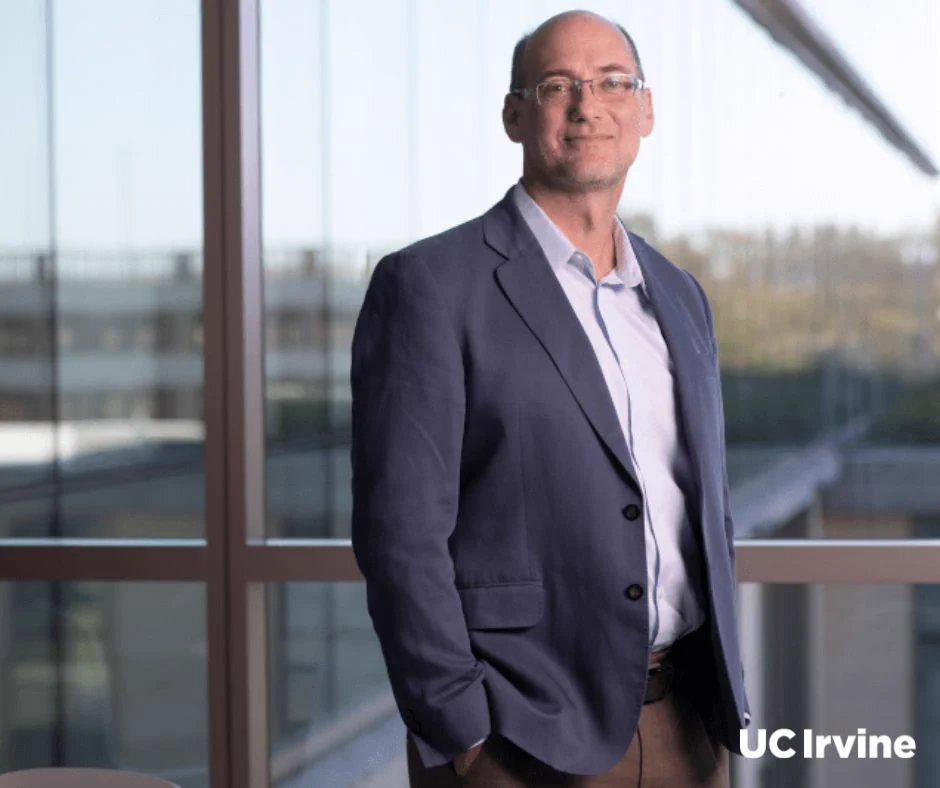 UC Irvine professor David Richardson poses in front of a glass window on the UC Irvine campus. He is wearing a blue blazer and collared shirt.