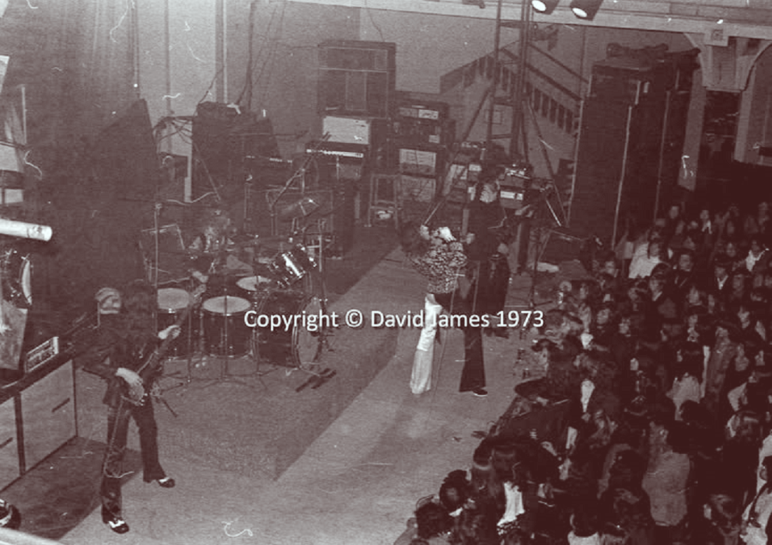 futatabirock1's tweet photo. 【何気ないある日のジョン】

＃JohnDeacon #Queen

1973年12月1日イギリスのサウスエンドで公演中のクイーン。
俯瞰で撮影された写真が不明瞭だけれど、拡大してみると初期の音響装置の配置や観客の様子が臨場感があって「情熱だけで突っ走ってたあの頃のクイーン」らしくって良い。
フレディのこの衣装の上下の組み合わせも珍しいのではないか。
Johnの足元には幾重にもコードがうねっている。

Kursaal, Southend, UK [01.12.1973]
引用元 QueenConcerts