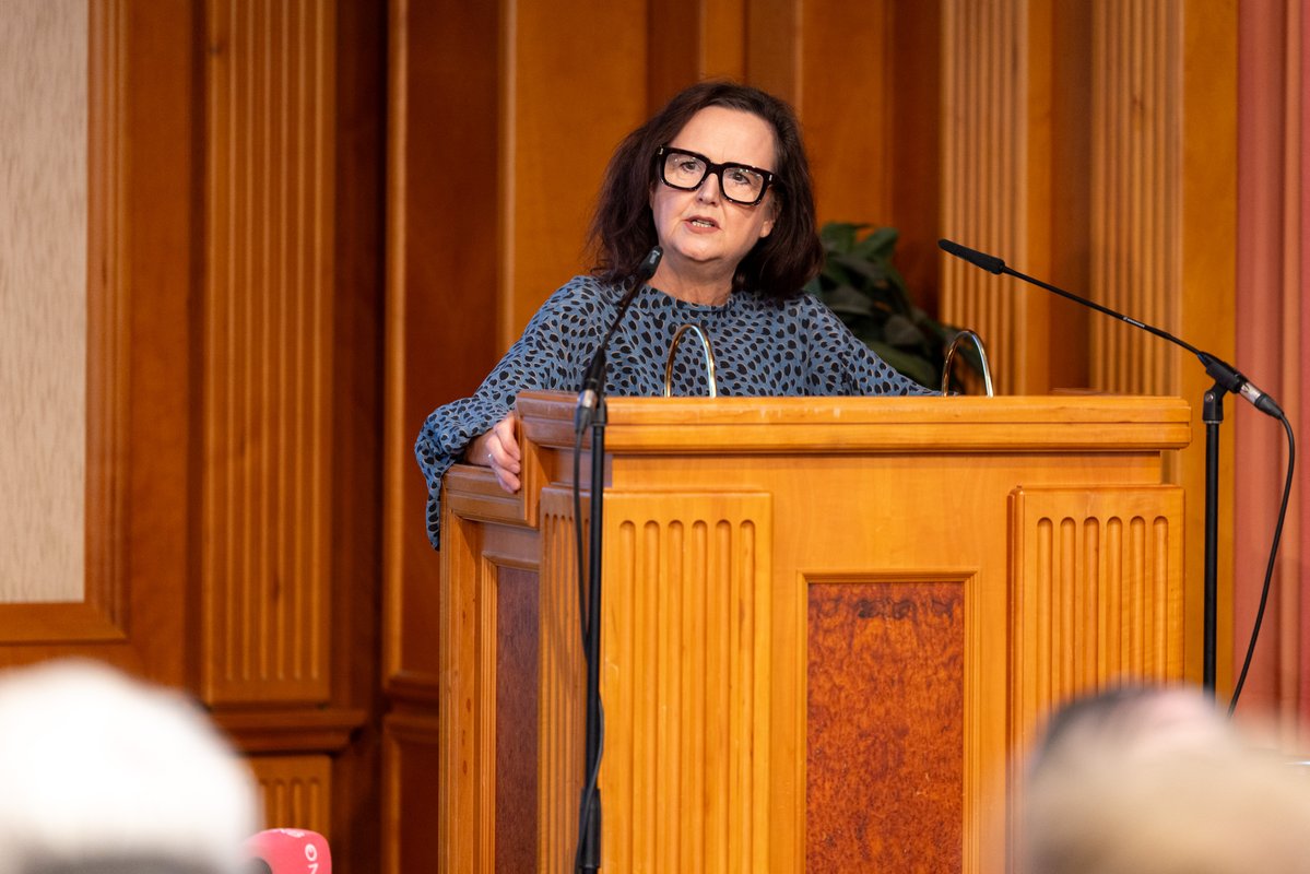 A person wearing glasses is standing at a podium delivering a speech.