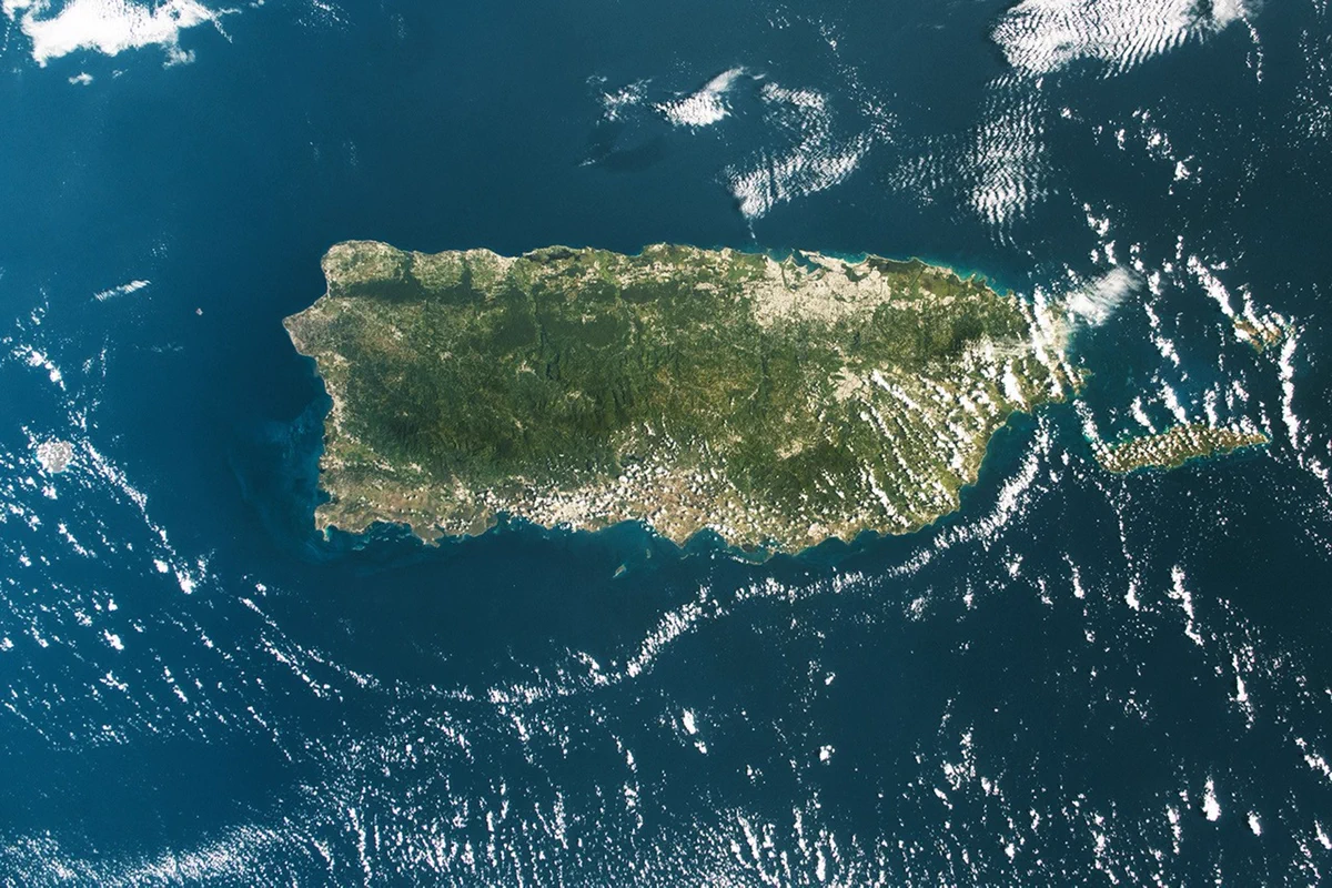 ISS astronaut photograph of Puerto Rico as seen from space. Most of the island is vivid green, with darker mountainous areas through the center and a noticeably browner, drier stretch along the southern coast. The island is surrounded by bright blue water and a few white clouds hang offshore.