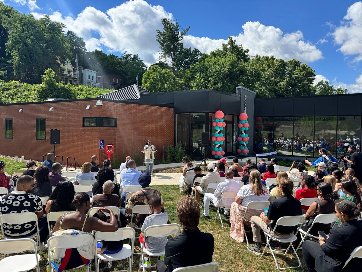 Community members gather outdoors for the grand opening of The Citizen Science Lab. Rows of attendees sit facing a speaker at a podium in front of the building, which is decorated with colorful balloon columns. The audience listens under a bright blue sky with trees and hillside homes visible in the background.