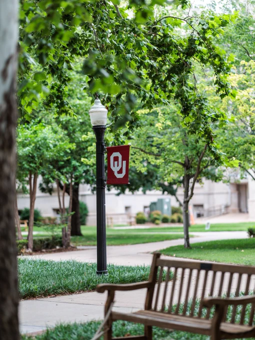 A red banner with the "OU" logo hangs on a lamp post in a lush, green park setting. There are trees, grassy areas, and a wooden bench nearby. A building is visible in the background.
