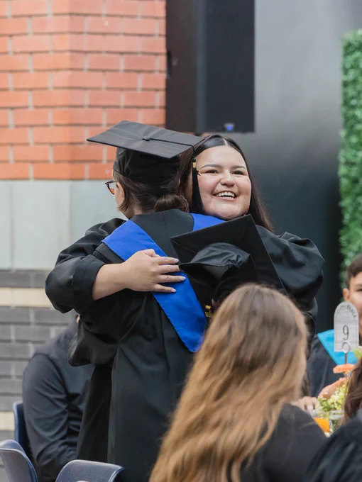 Two individuals in graduation caps and gowns are hugging, celebrating at a graduation event. They are indoors, surrounded by tables and seated guests. A brick wall is visible in the background.