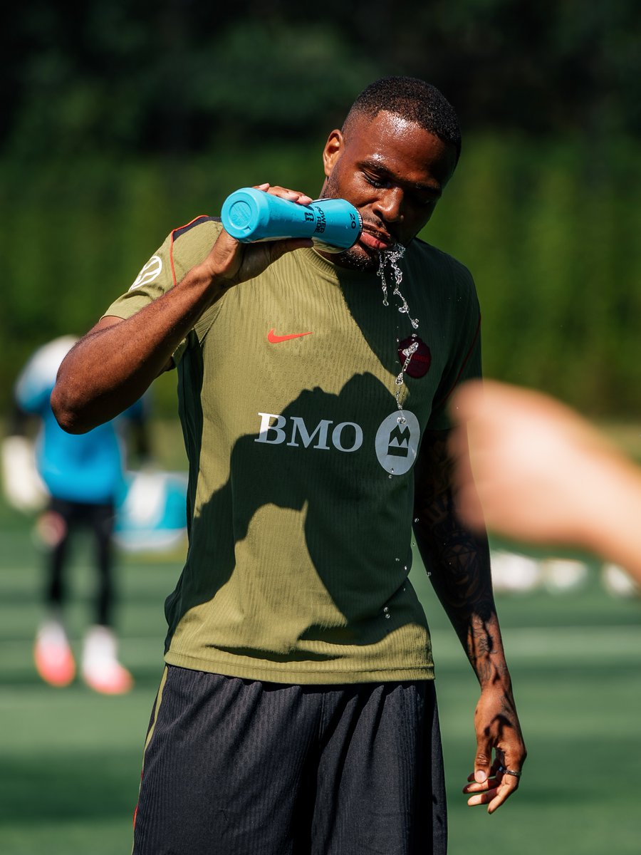 Cyle Larin drinking from a water bottle whilst training with Canada. He is wearing a brown training top with black shorts as water spills from his face.