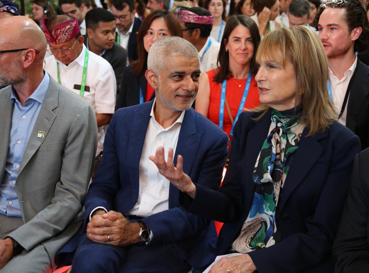 Mayor of London Sadiq Khan and a guest seated at a formal event talk in the front row, with a large audience behind them wearing conference badges.