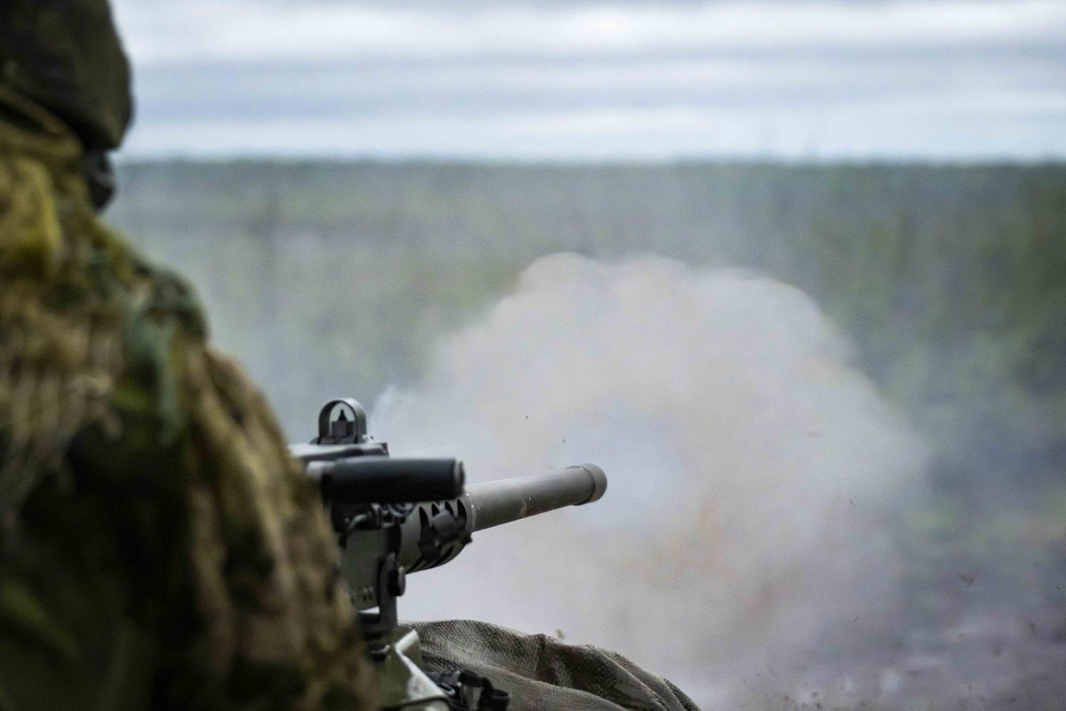 Soldat en tenue de camouflage tirant avec un fusil, de la fumée visible s’échappant du canon, dans un environnement extérieur.