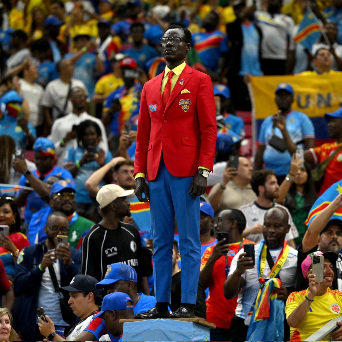chris_kabanga's tweet photo. Dans les tribunes, un supporter congolais en veste rouge se hisse au-dessus de la foule, bras tendus vers le ciel, comme pour offrir sa joie à tout un peuple. Lumumba Vea. https://t.co/RGmVPqRmkC