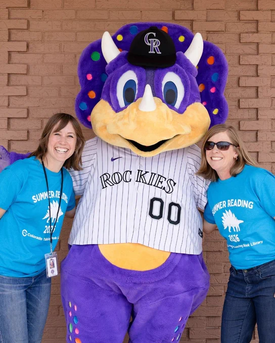 Two women in blue shirts hug the Rockies' mascot Dinger on either side of him in front of a brick wall.