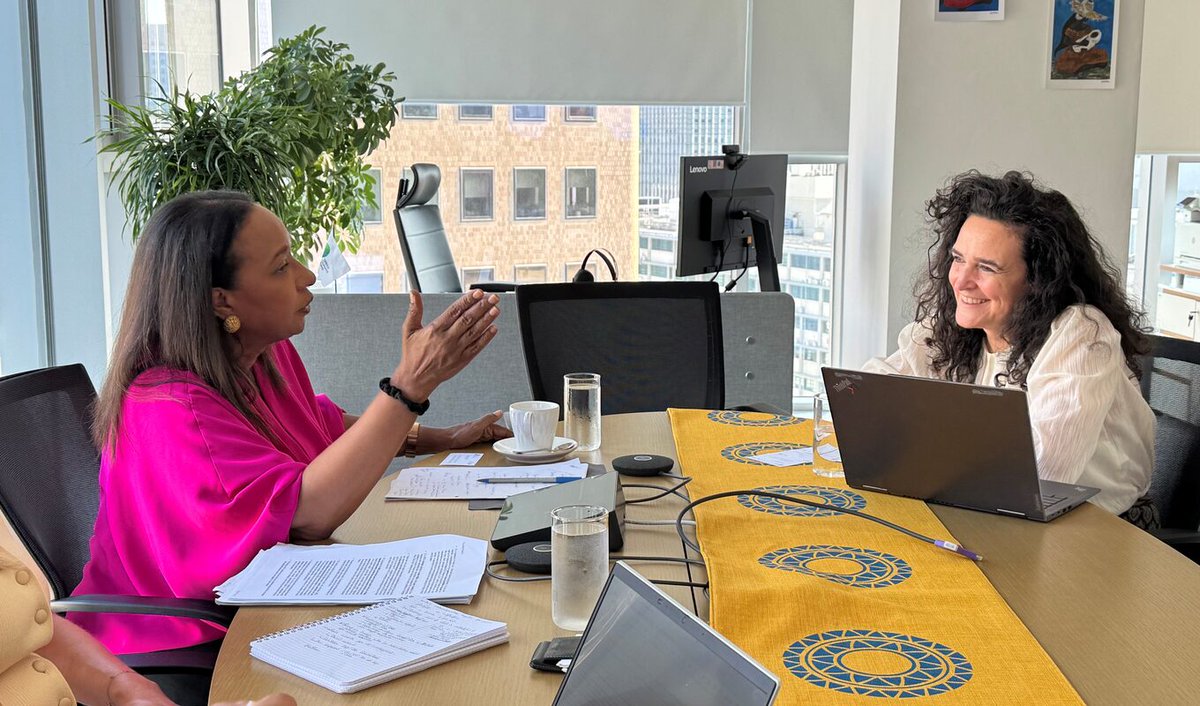 In a modern office setting, two people are engaged in discussion at a table with laptops and documents. The table has a yellow runner with circular patterns. Large windows in the background reveal a cityscape. A potted plant is visible to the left.