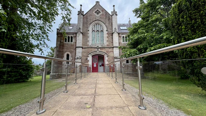 Gothic-style stone church with a large arched stained-glass window and red double doors, viewed from a tiled accessible walkway with stainless-steel and glass railings leading to the entrance, flanked by lawns and trees.