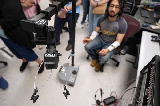 A man with dark hair and glasses sits in a chair and guides a robotic arm